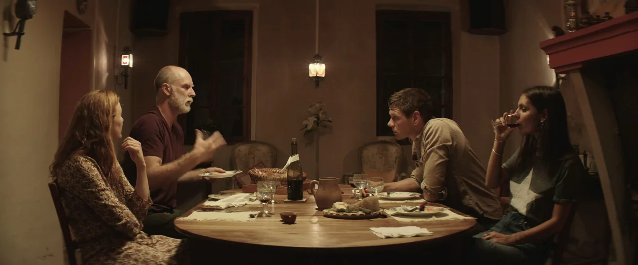 The Only One (2021) movie still: medium — A dimly lit room with a large wooden table and ornate chairs. Four individuals sit at the table with wine…