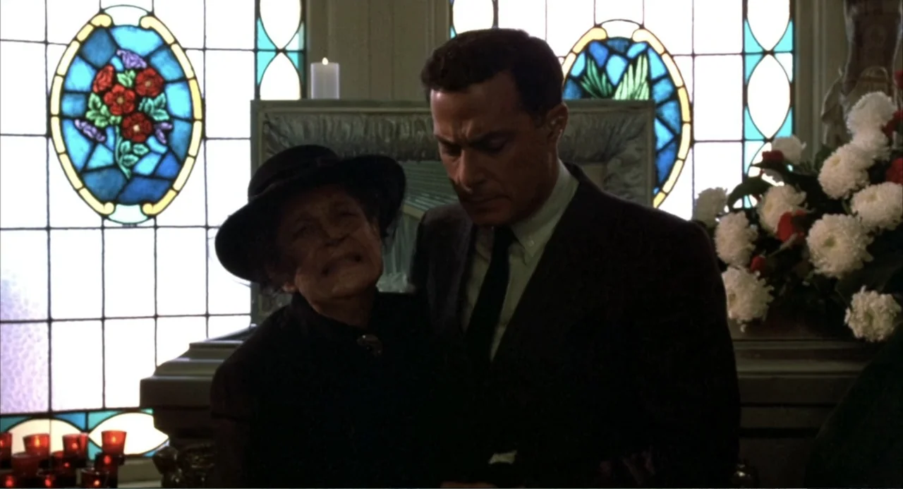 Murphy's Law (2009) movie still: medium — An interior space features tall stained glass windows behind a funeral display area. A man in a dark suit…