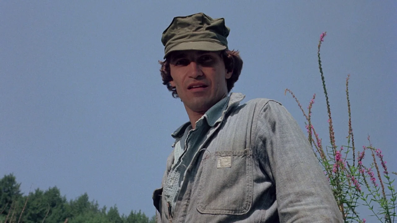 I Spit On Your Grave (1978) movie still: medium — A man in a utility cap stands in an open field against a clear sky. He wears a light denim jacket…