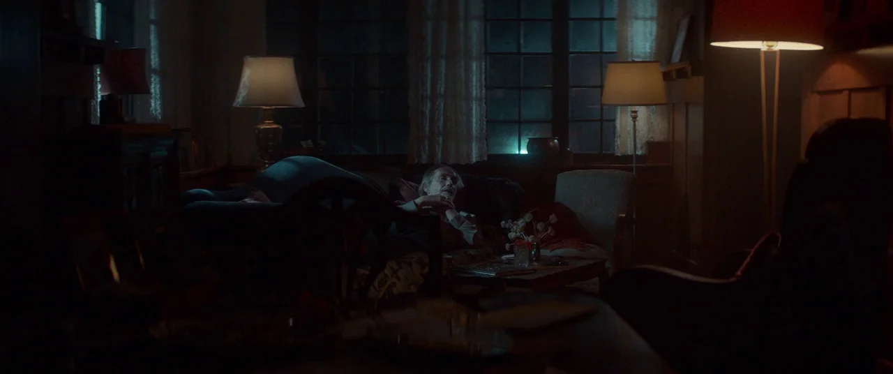 Woodshock (2017) movie still: wide — A dimly lit room contains multiple lamps, a sofa, and large windows at night. An elderly man rests on the couch…