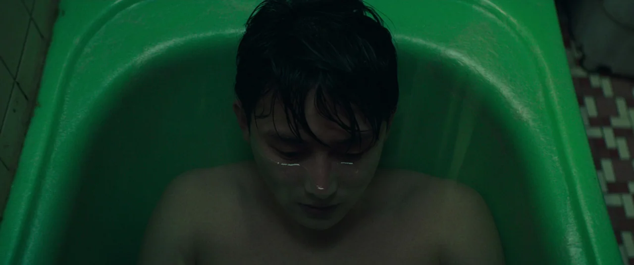 Beasts Clawing At Straws (2020) movie still: closeup — A person lies inside a bright green bathtub with tiled flooring visible to the side. The…