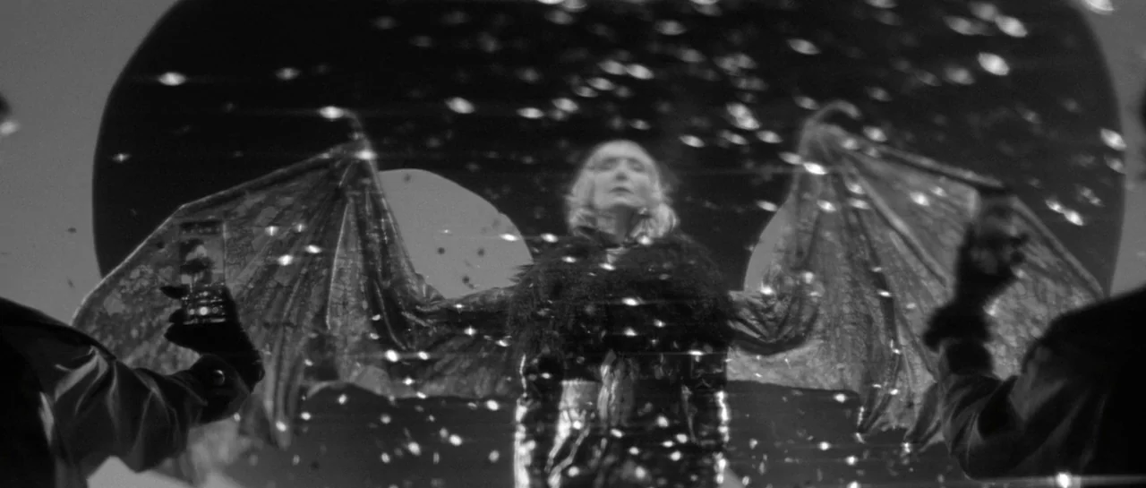 She Is Conann (2023) movie still: medium — A dark stage setting features a performer with large wings and a fur-trimmed coat. The figure stands…