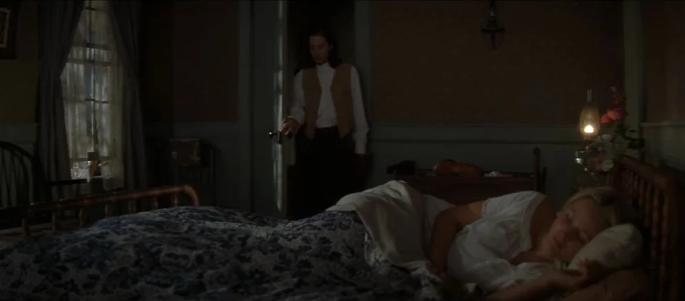 Ride With The Devil movie still: medium — A dimly lit room with dark walls, a patterned quilt, and a bedside oil lamp. A man stands in an open…