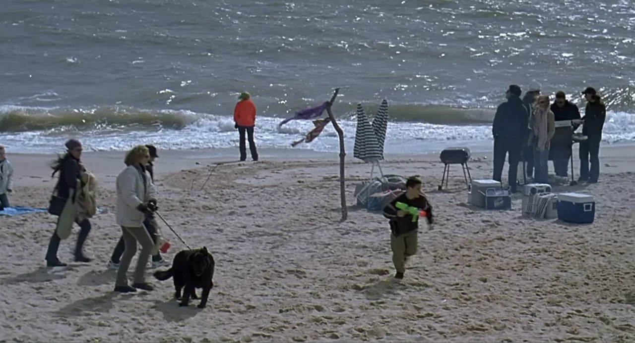 Eternal Sunshine of the Spotless Mind (2004) movie still: wide — A group of people gathers on a sandy shoreline with an ocean tide behind them. A…
