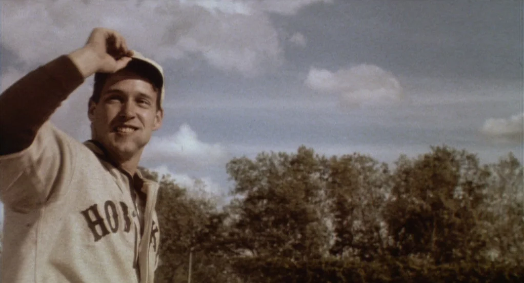 Eight Men Out (1988) movie still: medium — An outdoor baseball field with a treeline visible against a cloudy blue sky. A young man in a vintage…