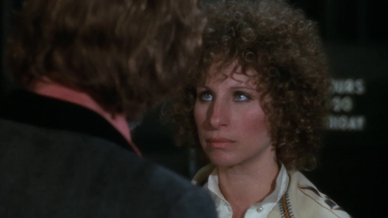 A Star Is Born (1976) movie still: medium — An interior space with dim lighting features a person with curly hair. The woman looks toward an unseen…