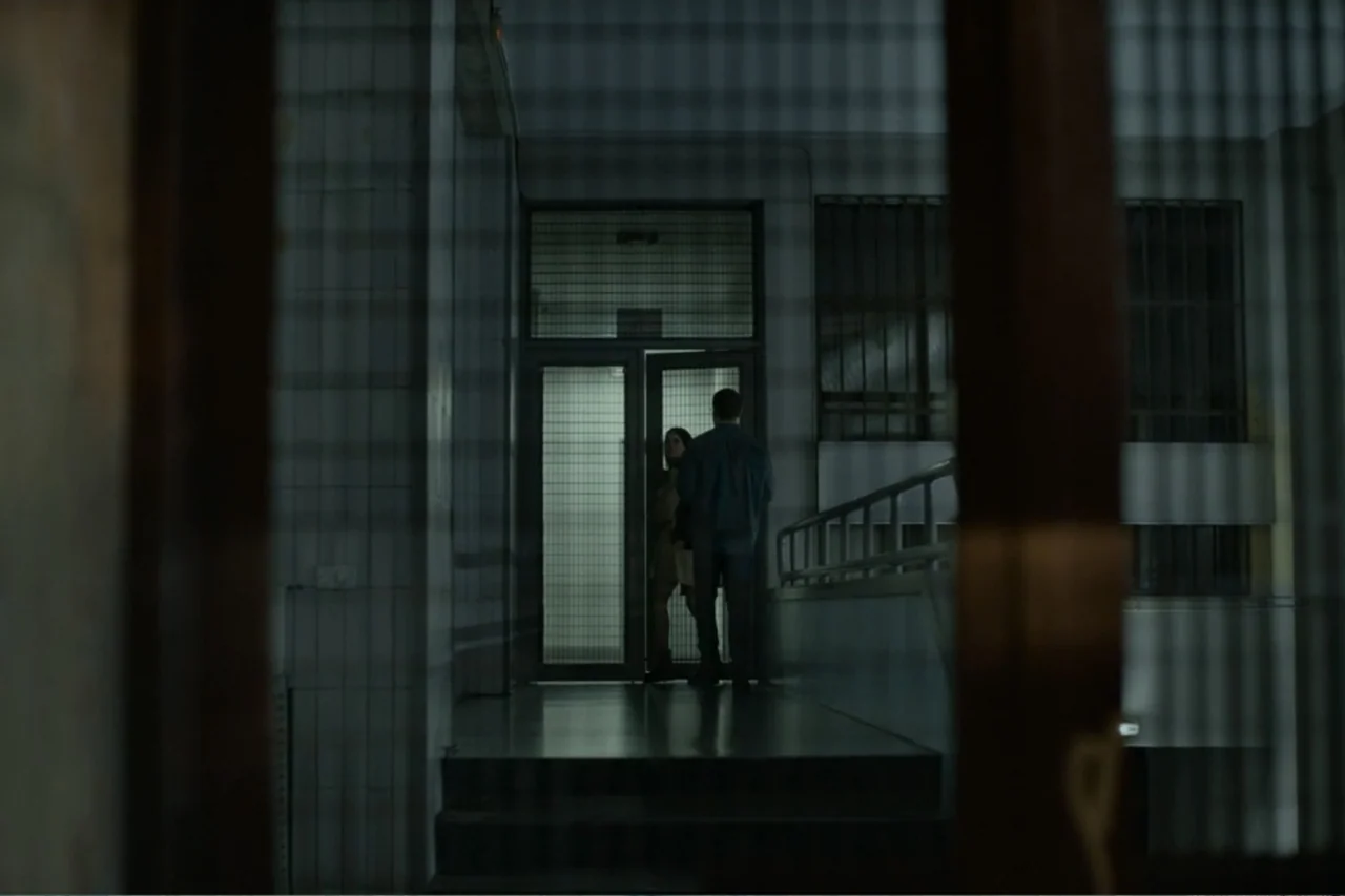 Cross the Line (2020) movie still: wide — A dimly lit, narrow corridor features tiled walls and a metal-grated glass doorway. A man and woman stand…