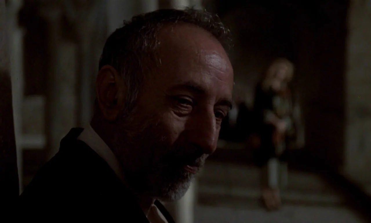 Nostalghia (1983) movie still: closeup — A dim interior with stone columns and a distant, blurred figure in the background. An elderly man stares…
