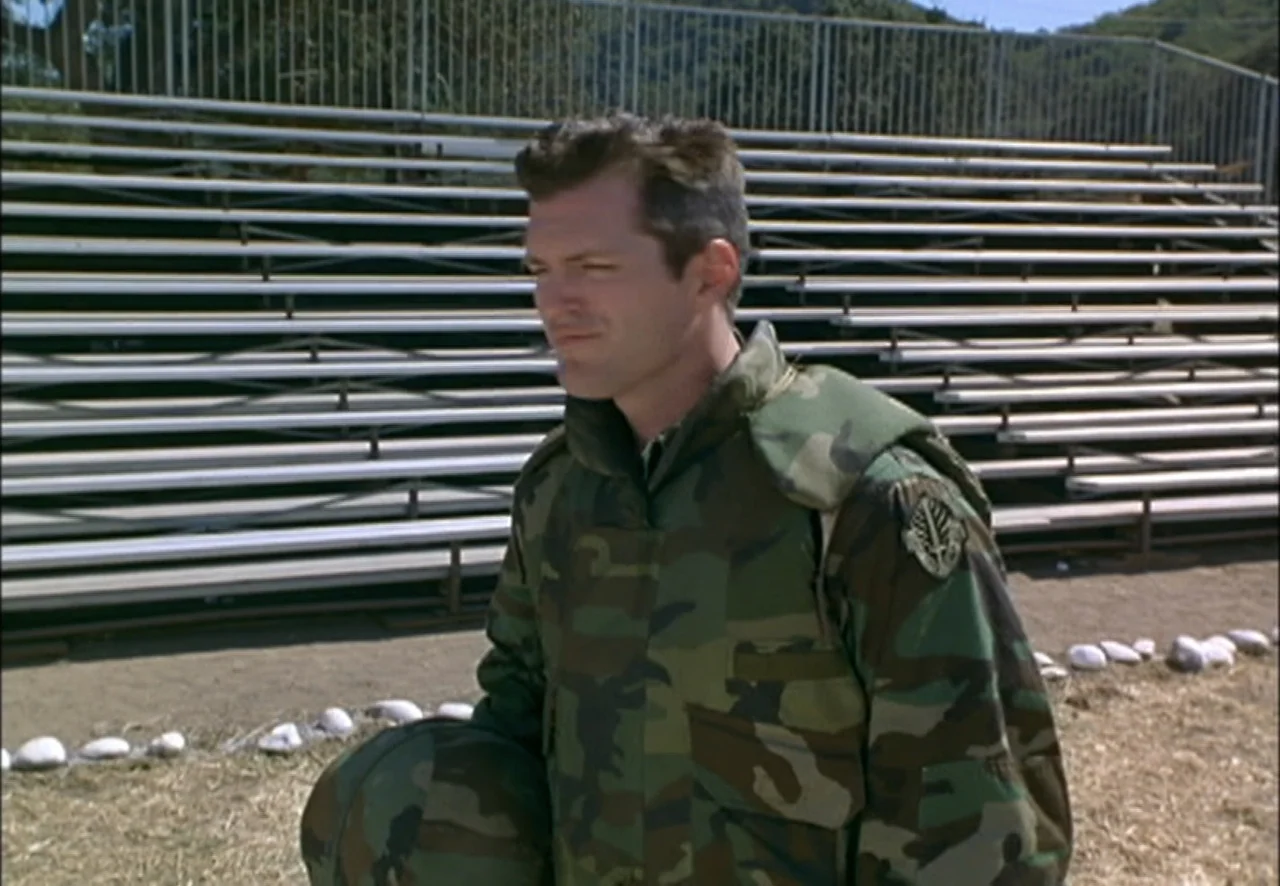 The Pentagon Wars (1998) movie still: medium — A man in military camouflage stands in front of empty metal stadium bleachers outside. He holds a…