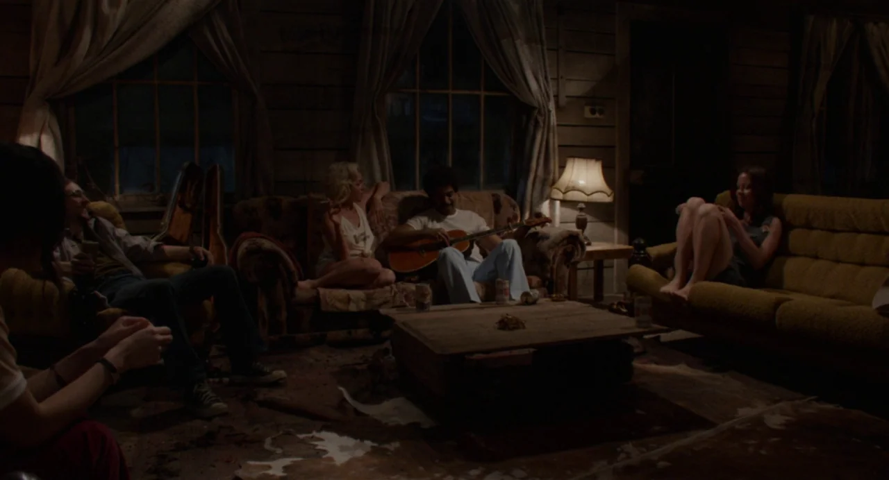 X (2022) movie still: wide — A dimly lit room with wooden walls and vintage 1970s furniture layout. A group of people lounge on sofas while one…