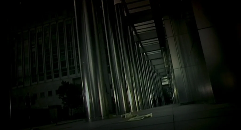 28 Days Later (2002) movie still: wide — A long covered walkway features rows of tall metallic columns under a modern building facade. The corridor…