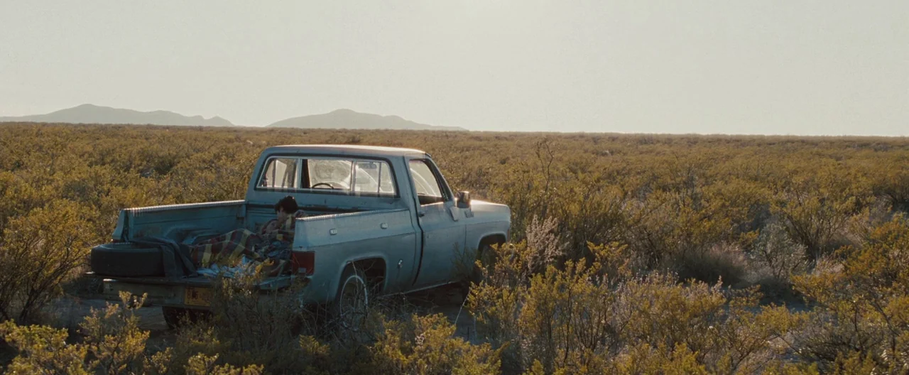 The Burning Plain (2008) movie still: wide — A silver pickup truck is parked in a vast, dry desert landscape with sparse bushes. A young man lies…