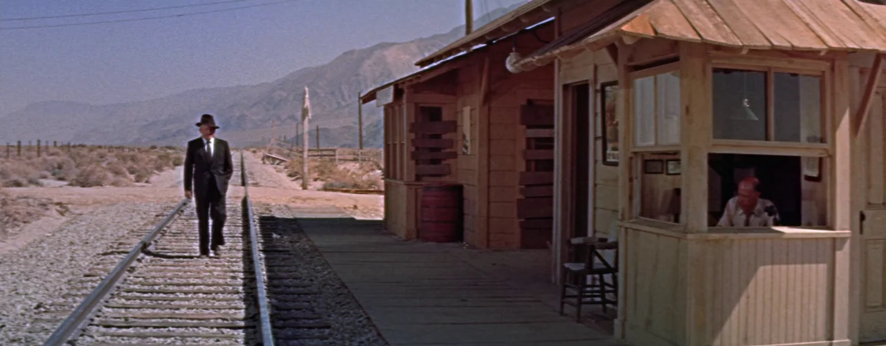Bad Day At Black Rock (1955) movie still: wide — A remote desert train station features wooden buildings and parallel railroad tracks under…