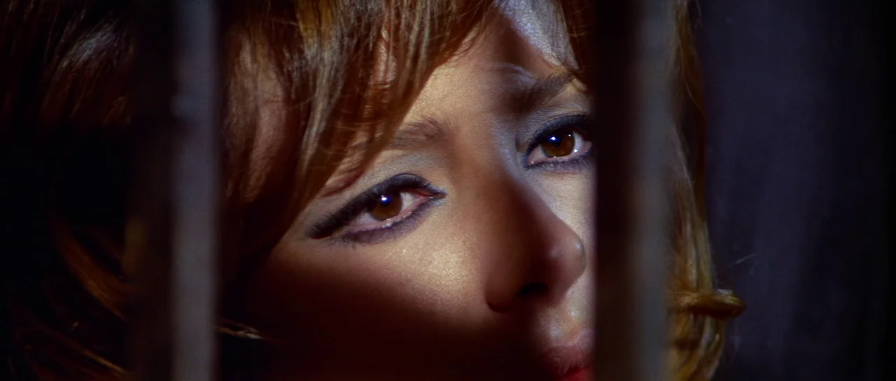 Night of the Blood Monster (1970) movie still: closeup — A close view shows a woman seen through the dark vertical bars of a barrier. She looks…