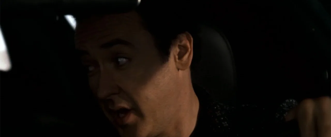 2012 (2009) movie still: closeup — The interior of a vehicle at night is dimly illuminated by external light. A man sitting in the driver's seat…