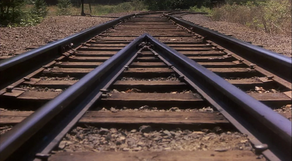 Stand By Me (1986) movie still: establishing — Railway tracks with wooden ties and crushed gravel extend into the distance. The rails converge at a…