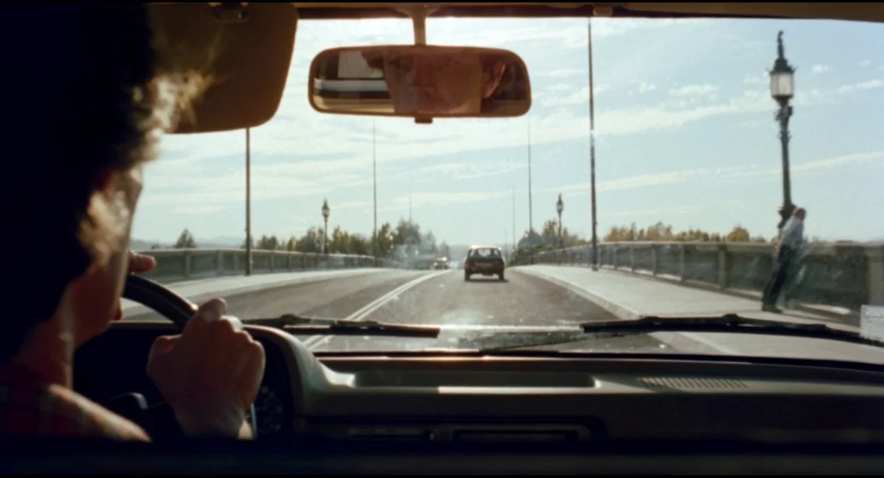 Olivia (1983) movie still: medium — A view from the interior of a car driving across a bridge during daytime. Another car is ahead on the road and a…