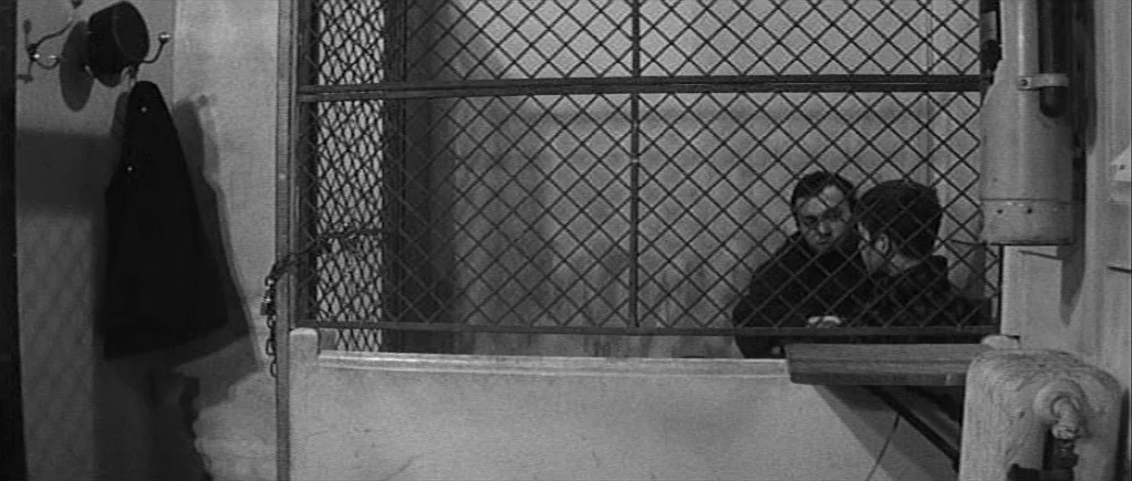 The 400 Blows (1959) movie still: medium — Two men sit inside an elevator cabin behind a heavy wire mesh partition. The small space includes a…