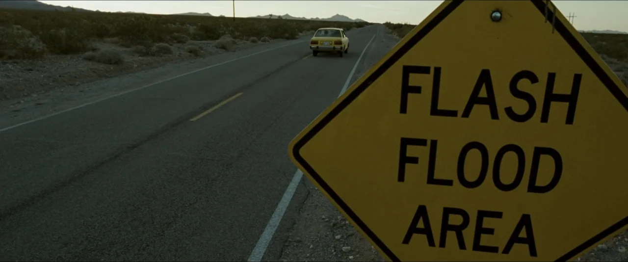 Into the Wild (2007) movie still: wide — A long asphalt road stretches through a flat desert under a clear sky. A yellow car moves away into the…