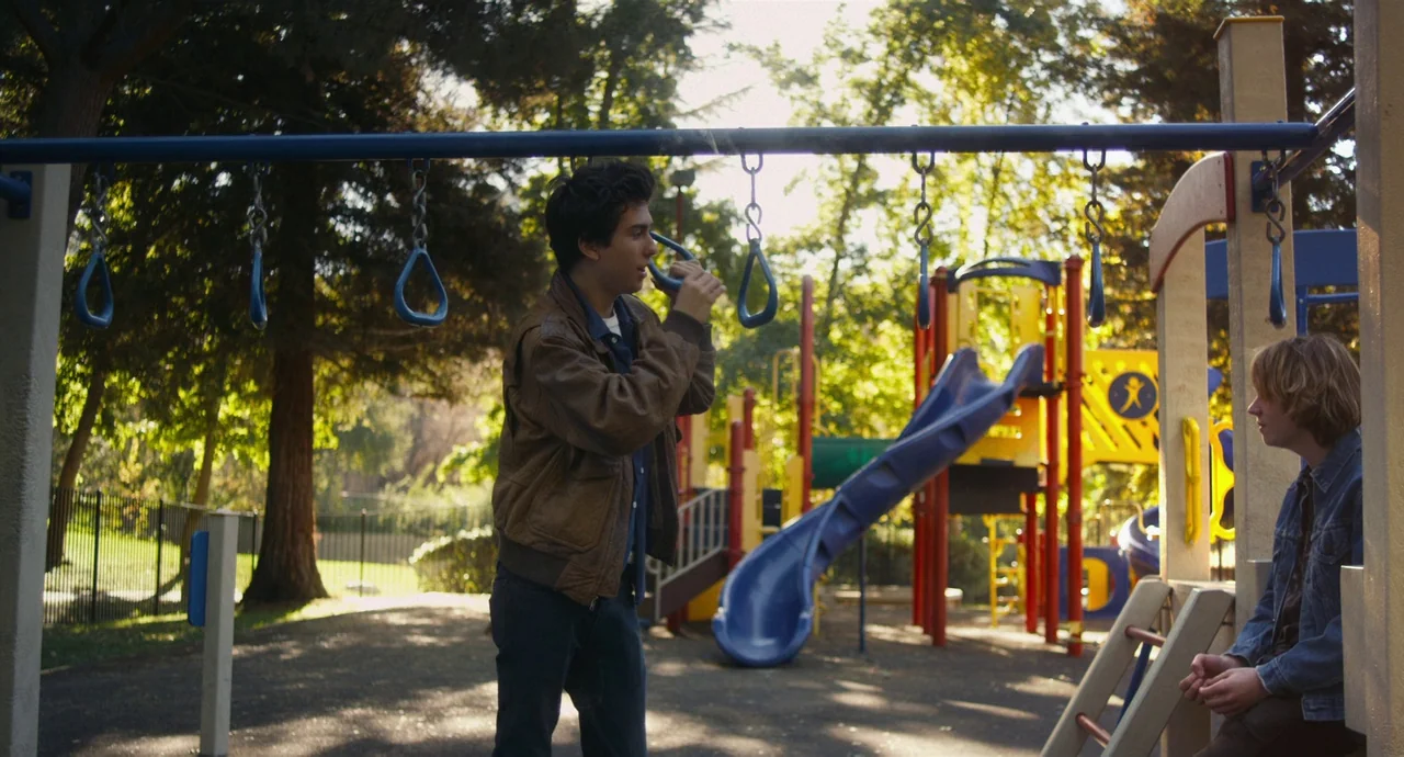 Palo Alto (2013) movie still: fullbody — A public park playground features colorful slides and metal monkey bar structures under trees. Two young…