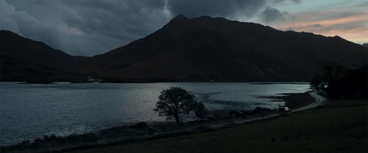 My Son (2021) movie still: establishing — A dark mountainous landscape overlooks a vast body of water at dusk. A single car drives along the road…