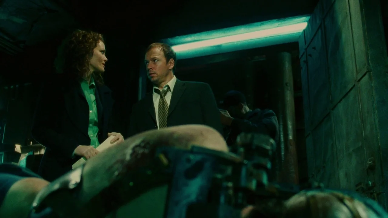 Saw II movie still: medium — A dimly lit morgue room with metallic surfaces and green-tinted fluorescent overhead lighting. A man and a woman in…