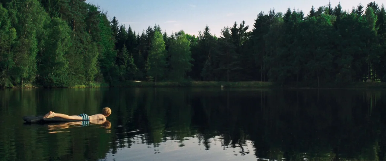 Goodnight Mommy (2014) movie still: wide — A dark calm lake is bordered by a dense wall of tall evergreen pine trees. A young boy lies on his…