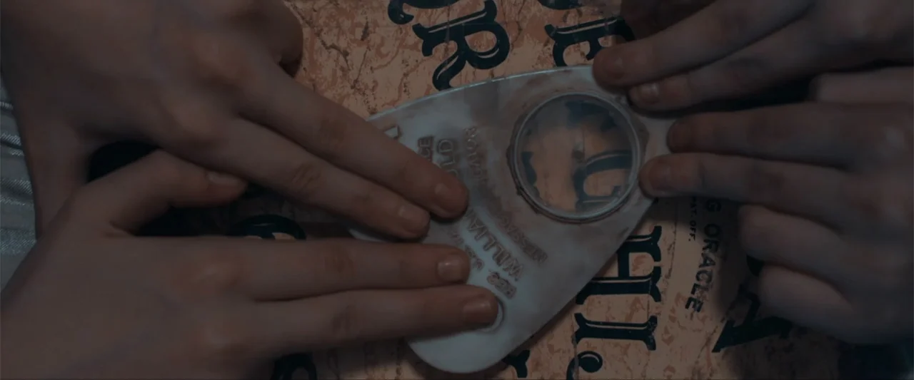 Anomaly (2006) movie still: closeup — A top-down view of a vintage spirit board with weathered tan surface texture. Four hands rest on a translucent…