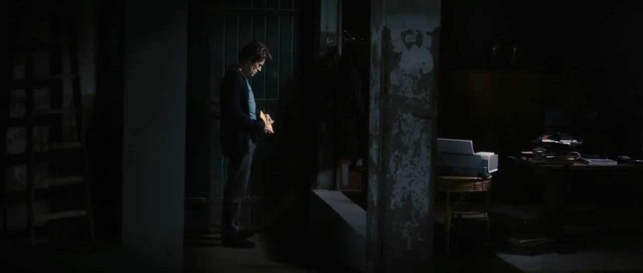 Antichrist (2009) movie still: medium — A dimly lit basement features concrete pillars and a wooden door with metal hinges. A man stands near a low…