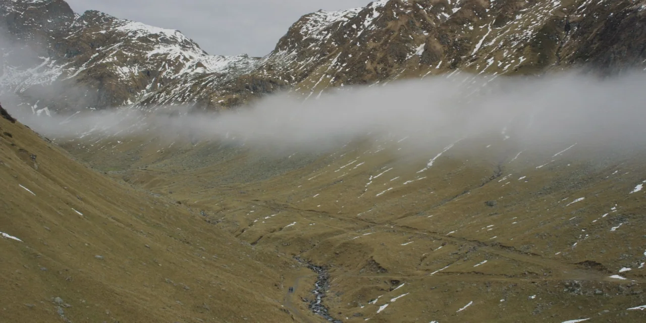 The Ritual (2017) movie still: wide — A wide mountain valley features grassy slopes with patches of remaining snow. A layer of low-hanging fog…