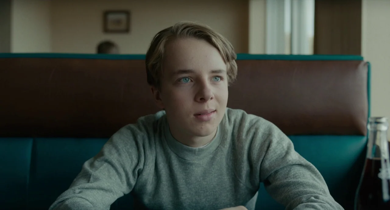 Wildlife (2018) movie still: medium — A diner booth has teal vinyl upholstery and a dark brown leather backrest. A teenage boy with blonde hair sits…
