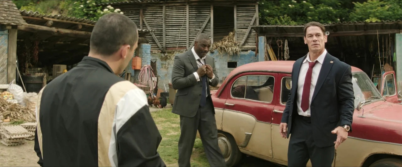 Heads of State (2025) movie still: medium — A rural courtyard features a weathered wooden barn and a two-tone classic sedan. Three men stand in…