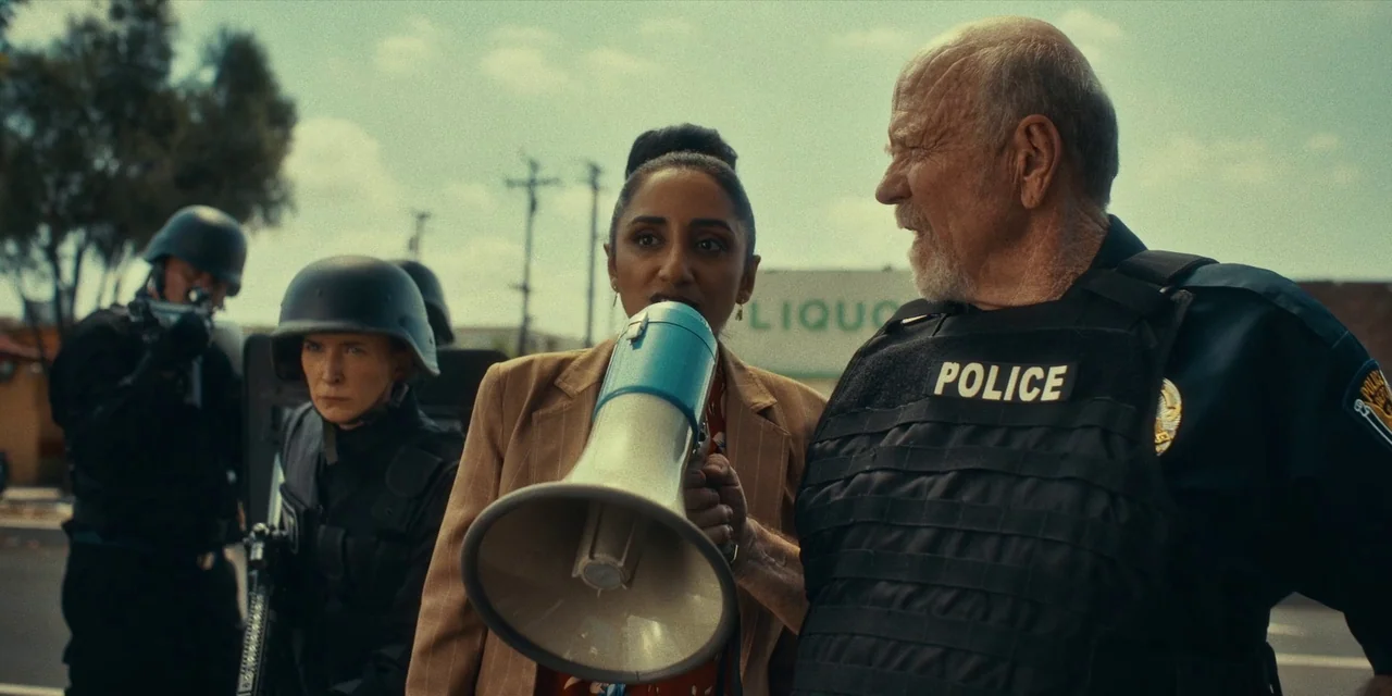 Destroy All Neighbours (2024) movie still: medium — A group of police officers and a civilian stand in a paved outdoor area. The woman holds a…