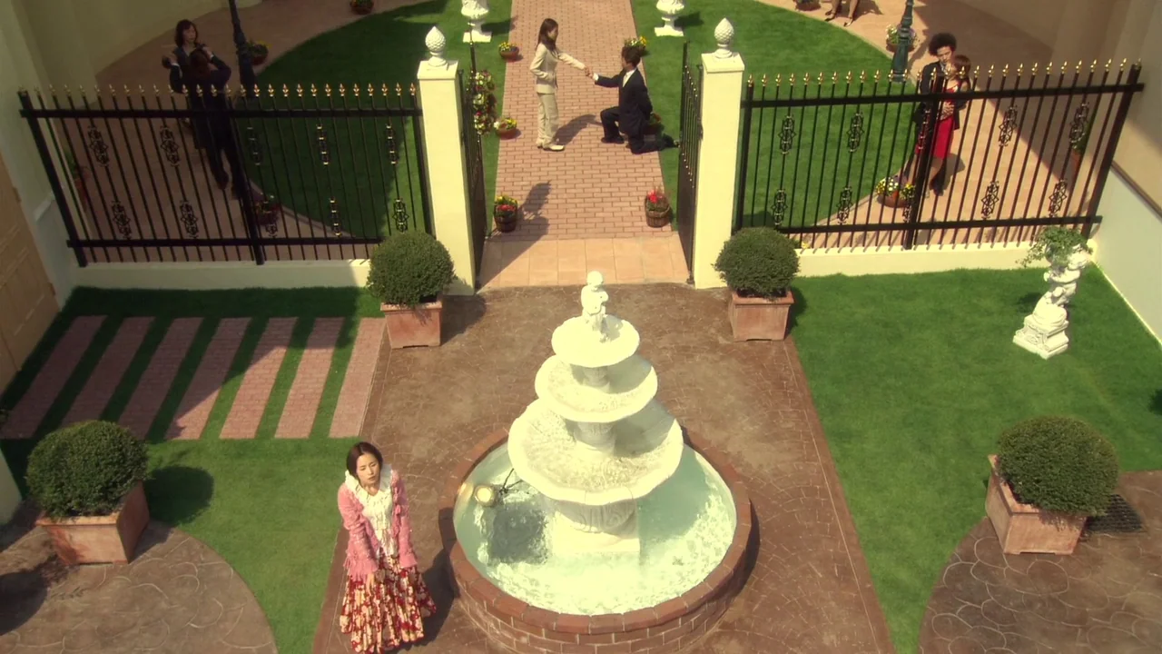 The Happiness of the Katakuris (2001) movie still: wide — A stone courtyard features a central tiered fountain, manicured lawn, and black metal…