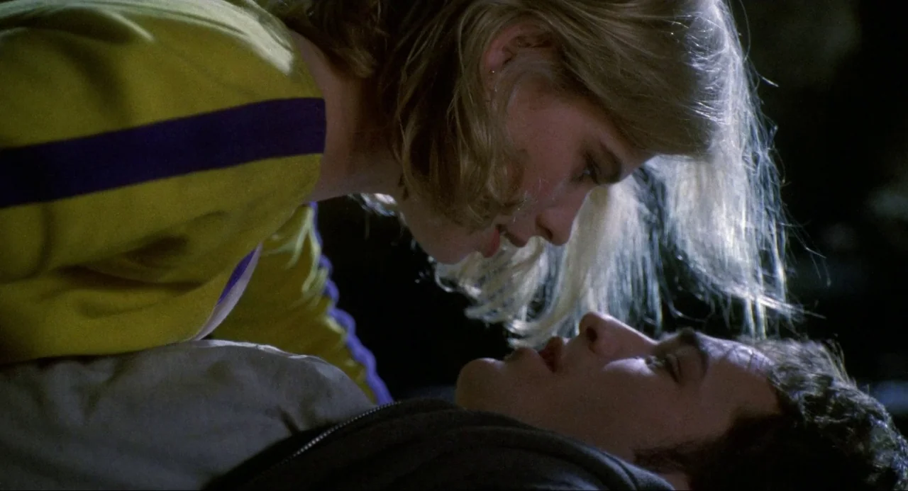 Buffy the Vampire Slayer (1992) movie still: closeup — A dark indoor space where two people lie face to face. A woman wearing yellow leans over a…