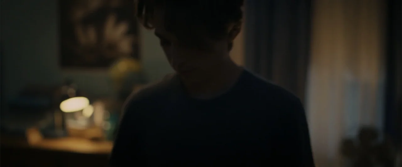 Chemical Hearts (2020) movie still: medium — A dark bedroom interior features a desk with a glowing lamp in the background. A young man stands in…
