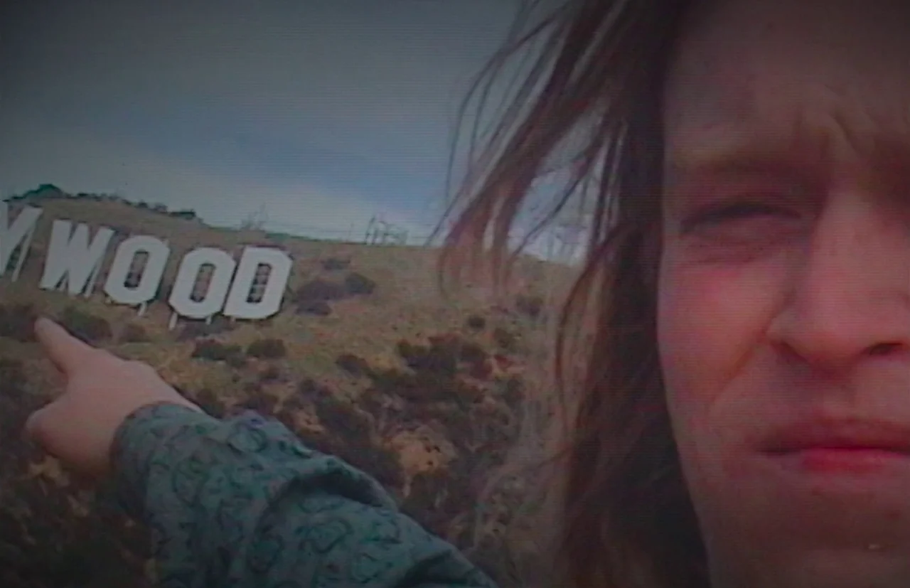 Nitram (2021) movie still: closeup — A dry, grassy hillside features large, white lettering spelling out a portion of a sign. A man with…