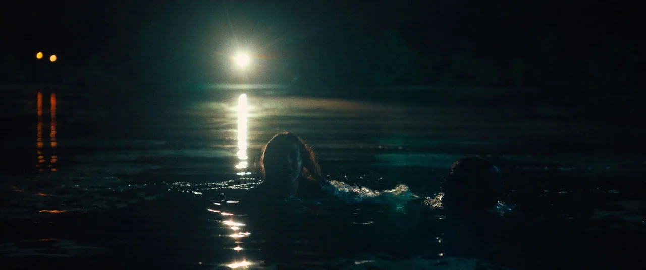 Dark Waters (2019) movie still: wide — Two individuals are swimming through dark, calm water at night. A single bright headlight shines from the…