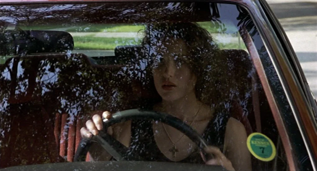 Sex, Lies And Videotape movie still: medium — A woman sits inside a car during the day with reflections of trees on the windshield. She holds the…