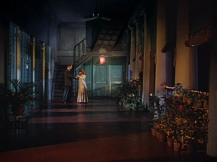 The River (1951) movie still: wide — A dimly lit covered veranda features structural columns, potted plants, and a dark wooden floor. A man in a…