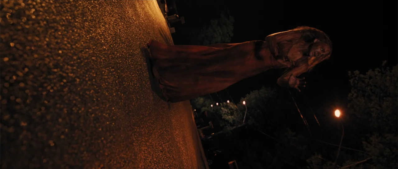 Carrie (2013) movie still: fullbody — An asphalt road at night illuminated by a single glowing orange street lamp nearby. A woman in a long dress…