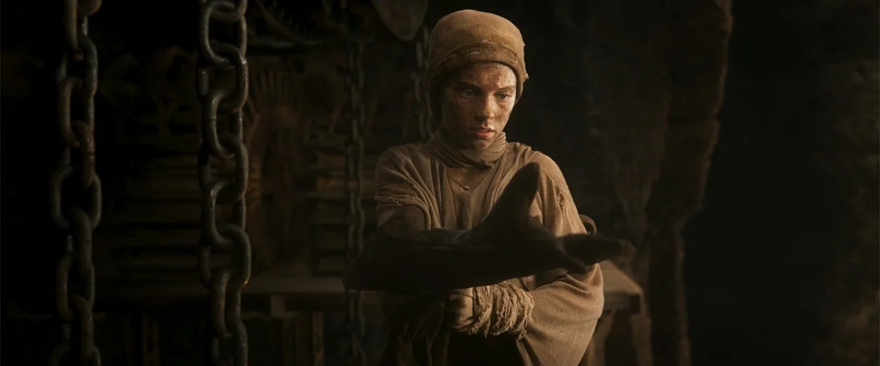 Furiosa: A Mad Max Saga (2024) movie still: medium — A dark smithy interior contains heavy iron chains hanging in the foreground. A teenage boy in…