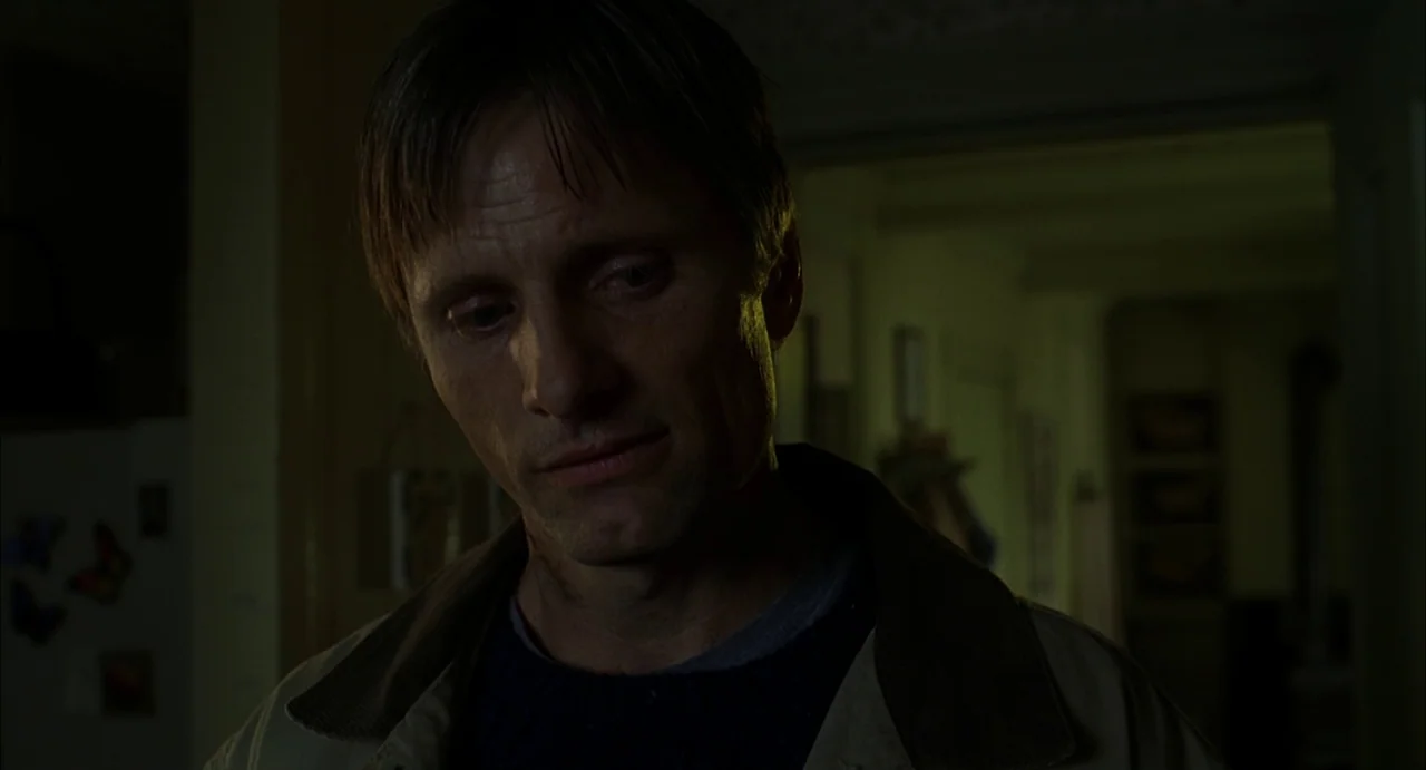 A History of Violence (2005) movie still: closeup — A dimly lit residential interior features a man standing in the foreground. He looks downward…