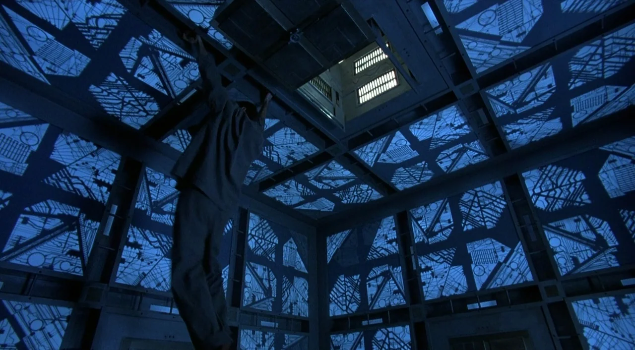 Cube (1997) movie still: wide — A dark, modular cube-shaped chamber features illuminated blue circuit patterns on all six surfaces. A person climbs…