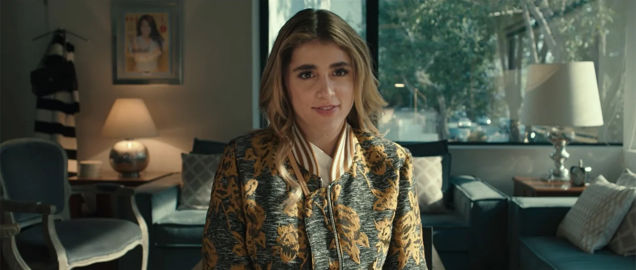 Cindy La Regia (2020) movie still: medium — A furnished interior features a sofa and lamps with a window revealing green foliage. A woman sits in…
