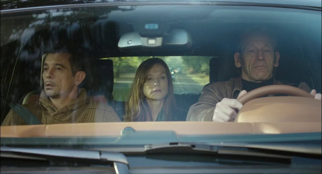 Abuse of Weakness (2013) movie still: medium — The interior of a vehicle on a bright day shows three adults sitting together. A man drives while a…