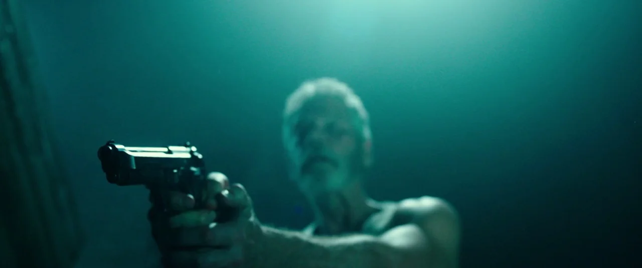 Don’t Breathe (2016) movie still: closeup — A man stands in an indeterminate dark space filled with green-tinted shadows. He extends a handgun…