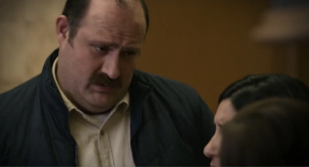 La posada (2023) movie still: medium — An interior space with beige walls providing a neutral backdrop for two people. A man wearing a vest observes…