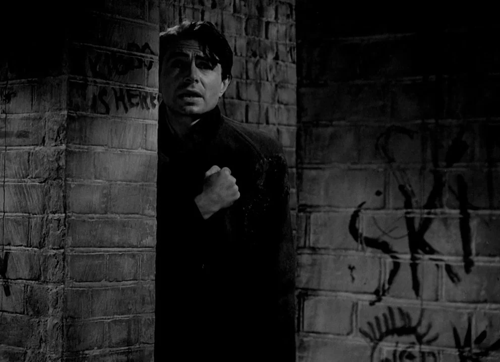 Odd Man Out (1947) movie still: medium — A man peers from behind the corner of a brick wall marked by spray-painted graffiti. He holds one hand…