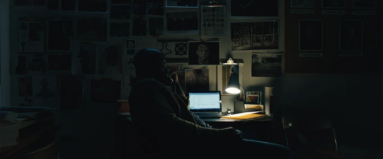 Skin (2009) movie still: medium — A dimly lit home office wall is covered with numerous photographs and printed documents. A man sits at a desk…
